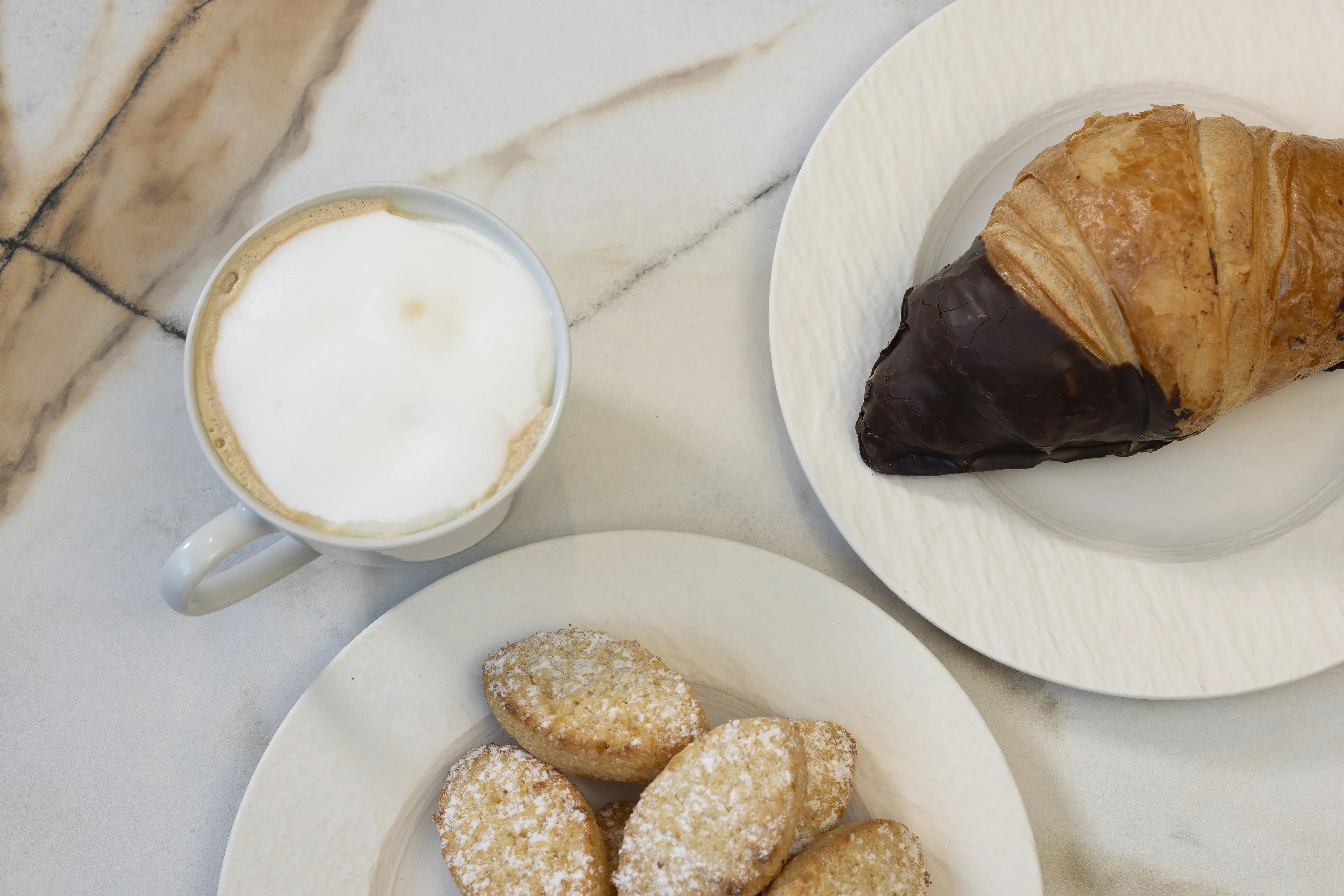 Tasse Cappuccino, Schokocroissant und kleine Kekse auf weißen Tellern.