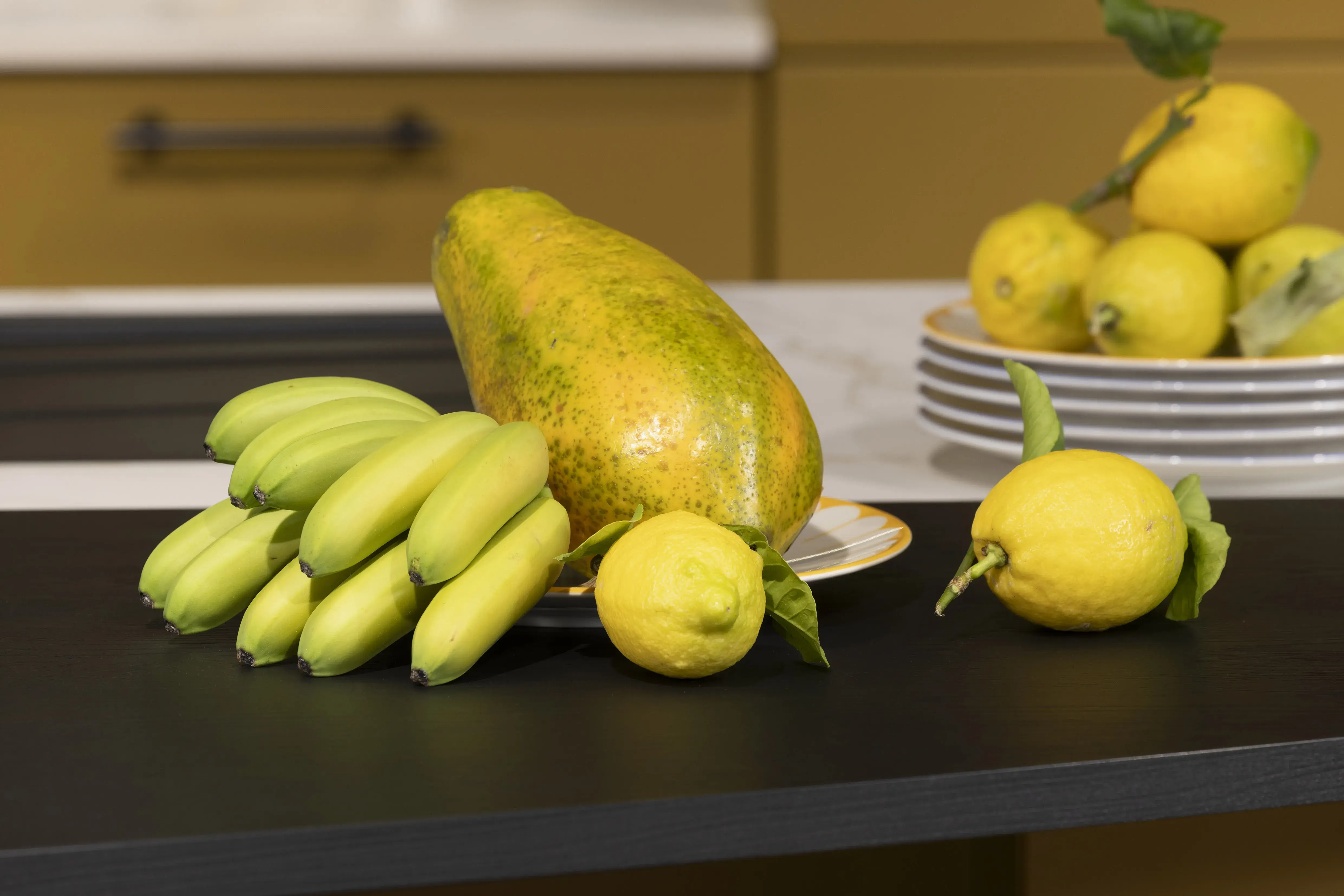 Papaya, Mini-Bananen und Zitronen liegen auf einem Tisch, im Hintergrund Teller mit mehr Zitronen.