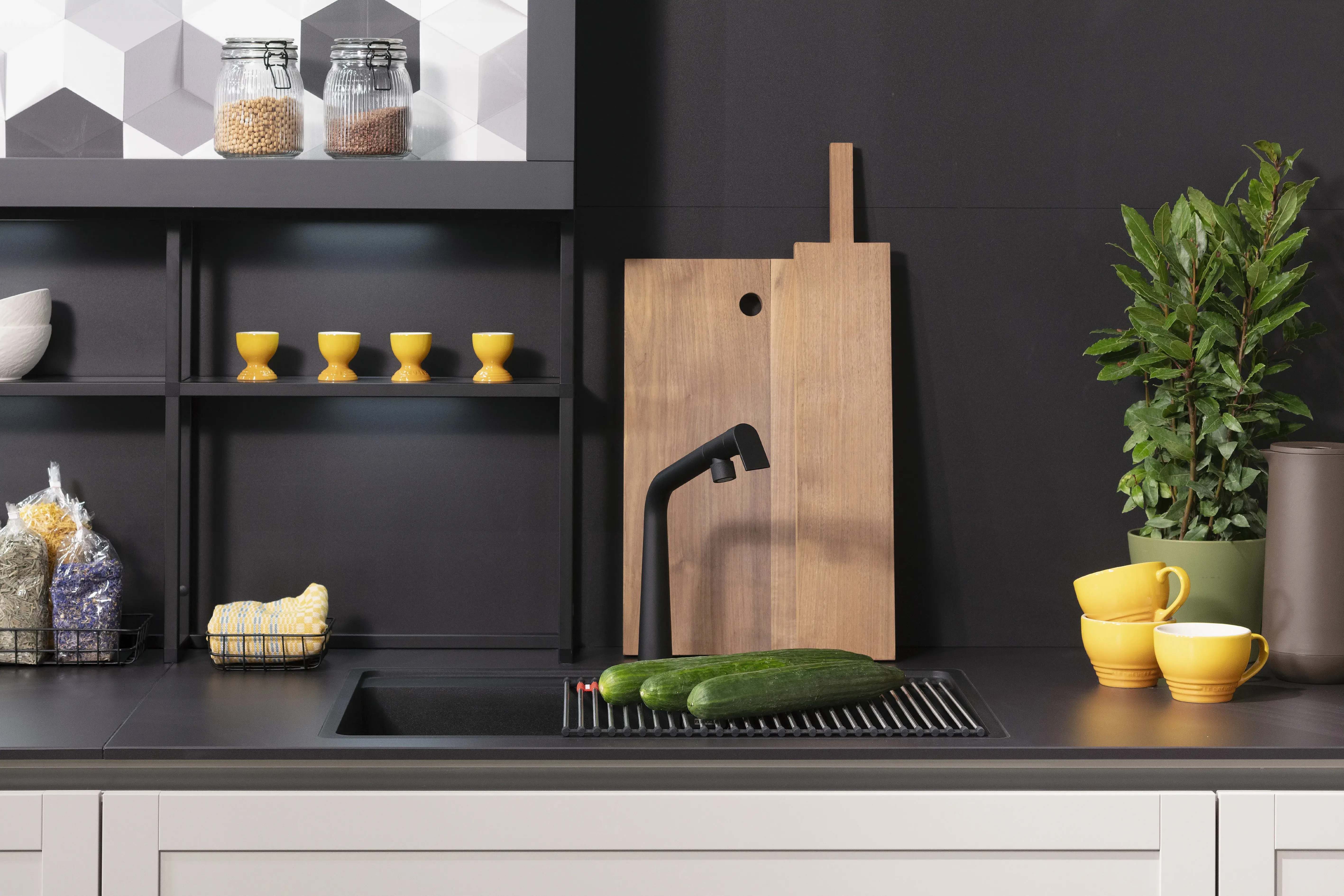 Moderne Küche mit schwarzer Wand, Holzbrettern, gelbem Geschirr und zwei Gurken am Waschbecken.